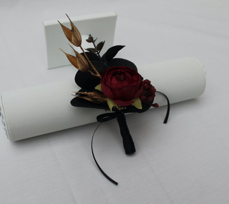 Pu&ograve; includere: Un boutonniere per un matrimonio o un evento speciale con una rosa bordeaux, dettagli dorati e fogliame nero, legato con un nastro nero.