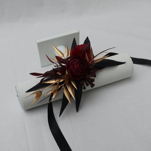 Pu&ograve; includere: Un arrangiamento floreale di colore bordeaux e oro con nastro nero, avvolto attorno a un cilindro bianco.
