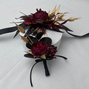 Pu&ograve; includere: Un arrangiamento floreale di colore bordeaux e oro con una scatola bianca. I fiori sono disposti in un bouquet con un nastro nero e sono legati insieme con un fiocco.