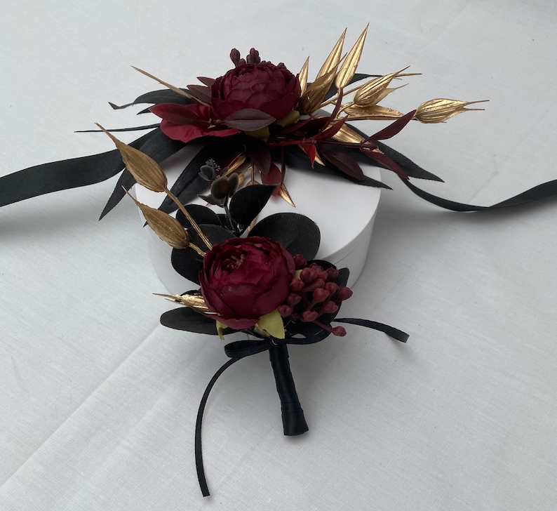 Pu&ograve; includere: Un arrangiamento floreale di colore bordeaux e oro con una scatola bianca. I fiori sono disposti in un bouquet con un nastro nero e sono legati insieme con un fiocco.