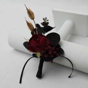 Pu&ograve; includere: Una rosa borgogna con foglie di eucalipto nere ed erbe secche dorate, legate con un nastro nero, &egrave; esposta su un supporto bianco. Si tratta di una boutonni&egrave;re, un piccolo arrangiamento floreale da indossare sul risvolto.