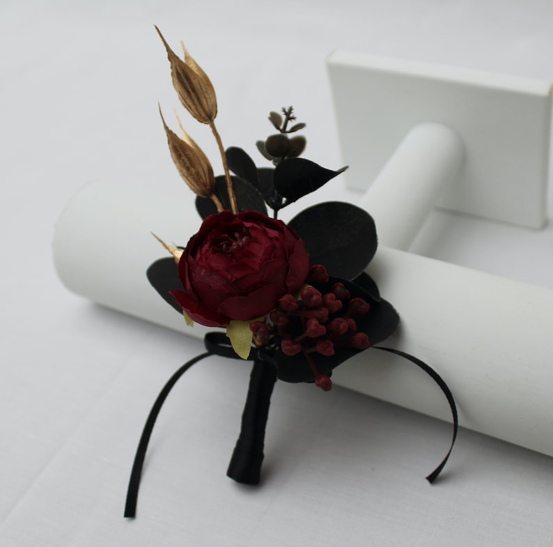 Pu&ograve; includere: Una rosa borgogna con foglie di eucalipto nere ed erbe secche dorate, legate con un nastro nero, &egrave; esposta su un supporto bianco. Si tratta di una boutonni&egrave;re, un piccolo arrangiamento floreale da indossare sul risvolto.