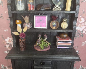 May include: Miniature black hutch with shelves displaying various items. Includes glass bottles with cork stoppers, a crystal ball, a mushroom figurine, a framed pink image, and small decorative perfume bottles. The bottom has drawers and a cabinet.