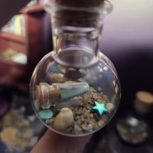 May include: A small, clear, spherical glass bottle with a cork stopper containing sand, small light blue stones, a tiny metallic blue star, and a miniature glass bottle with a cork stopper inside, creating a layered, textured, and decorative display.