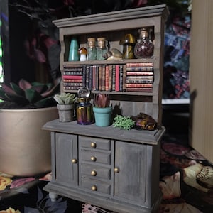 Op de afbeelding: Een miniatuur grijze houten kast met een boekenplank, lades en kastdeuren. De planken bevatten kleine boeken, glazen flessen en decoratieve items. Kleine planten in potten en een kleurrijke iriserende fles staan bovenop.