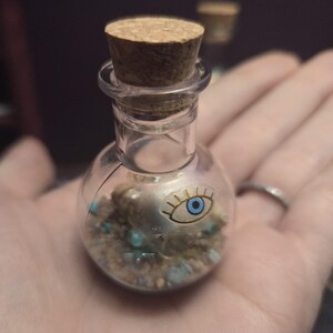 May include: A small, clear glass bottle with a cork stopper containing a layer of sand, small blue stones, and a silver-toned charm shaped like an eye with a blue iris. The bottle has a rounded base and a narrow neck, showcasing the contents inside.