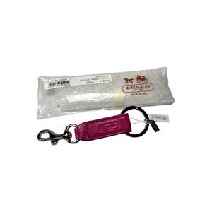 May include: A pink leather key chain with a silver key ring and a silver clip. The keychain has the word "COACH" embossed on it. The keychain is packaged in a clear plastic bag with a white tag that says "COACH EST. 1941 NEW YORK".