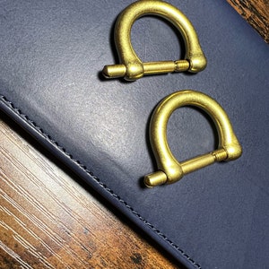 May include: Two brass-colored D-ring shackles with screw pins. The shackles are positioned on a dark blue surface, with a wooden surface visible in the background. These hardware components are likely used for various crafting or industrial applications.