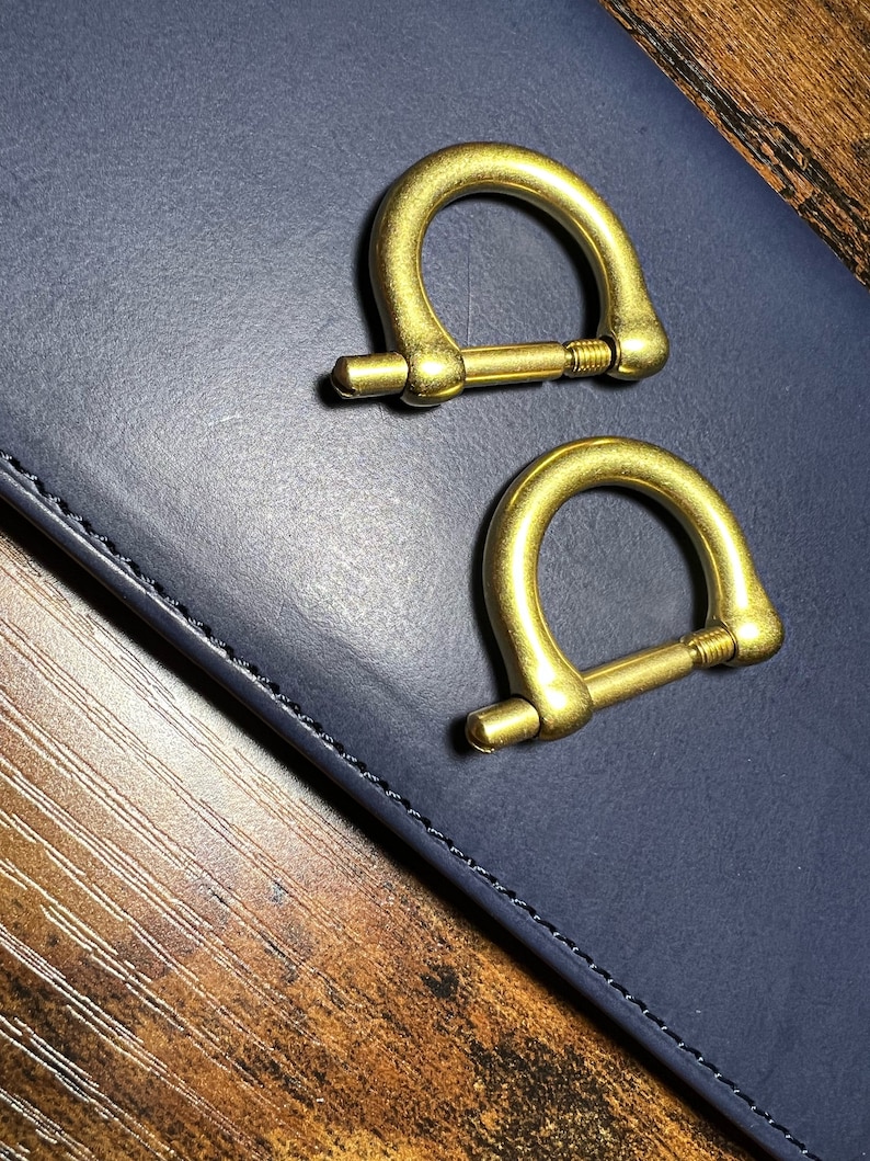 May include: Two brass-colored D-ring shackles with screw pins. The shackles are positioned on a dark blue surface, with a wooden surface visible in the background. These hardware components are likely used for various crafting or industrial applications.