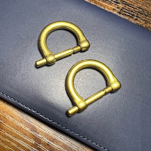 May include: Two brass-colored D-ring shackles with screw pins rest on a dark blue leather surface. The shackles have a curved, semi-circular shape with a threaded pin for secure closure. The background is a wooden surface.