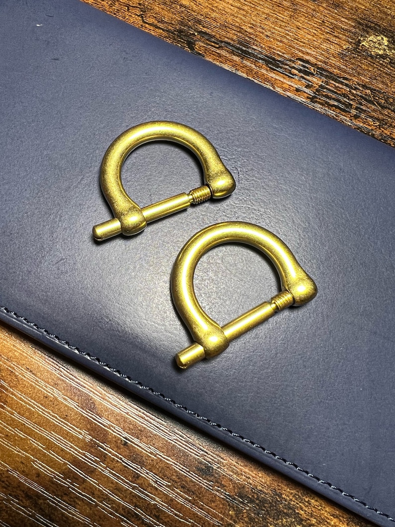 May include: Two brass-colored D-ring shackles with screw pins rest on a dark blue leather surface. The shackles have a curved, semi-circular shape with a threaded pin for secure closure. The background is a wooden surface.