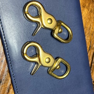 May include: Two gold-colored metal clasps with a swivel hook and a D-ring. The clasps are on a dark blue leather surface.