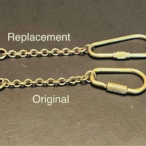 May include: Two gold-colored keychains, one labeled "Replacement" and the other "Original". Each keychain features a chain and a carabiner-style clasp. The clasps have different textured designs. The background is a dark, solid color.