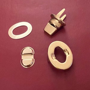 May include: Four gold-toned metal pieces for a purse or bag closure. The pieces include a round ring, a small oval with two holes, a larger oval with two holes, and a hook with two prongs.
