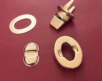 Könnte beinhalten: Vier goldfarbene Metallteile für einen Taschen- oder Handtaschenverschluss. Die Teile umfassen einen runden Ring, ein kleines Oval mit zwei Löchern, ein größeres Oval mit zwei Löchern und einen Haken mit zwei Zinken.