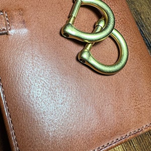 May include: A close-up shot of a brown leather wallet with a gold-colored metal clasp. The clasp is in the shape of two interlocking D-rings. The leather has a textured appearance and is stitched around the edges.