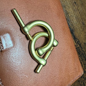 May include: A brass-colored metal clasp with two curved sections and a screw mechanism. The clasp is positioned on a brown leather surface, with a wooden surface visible in the background.