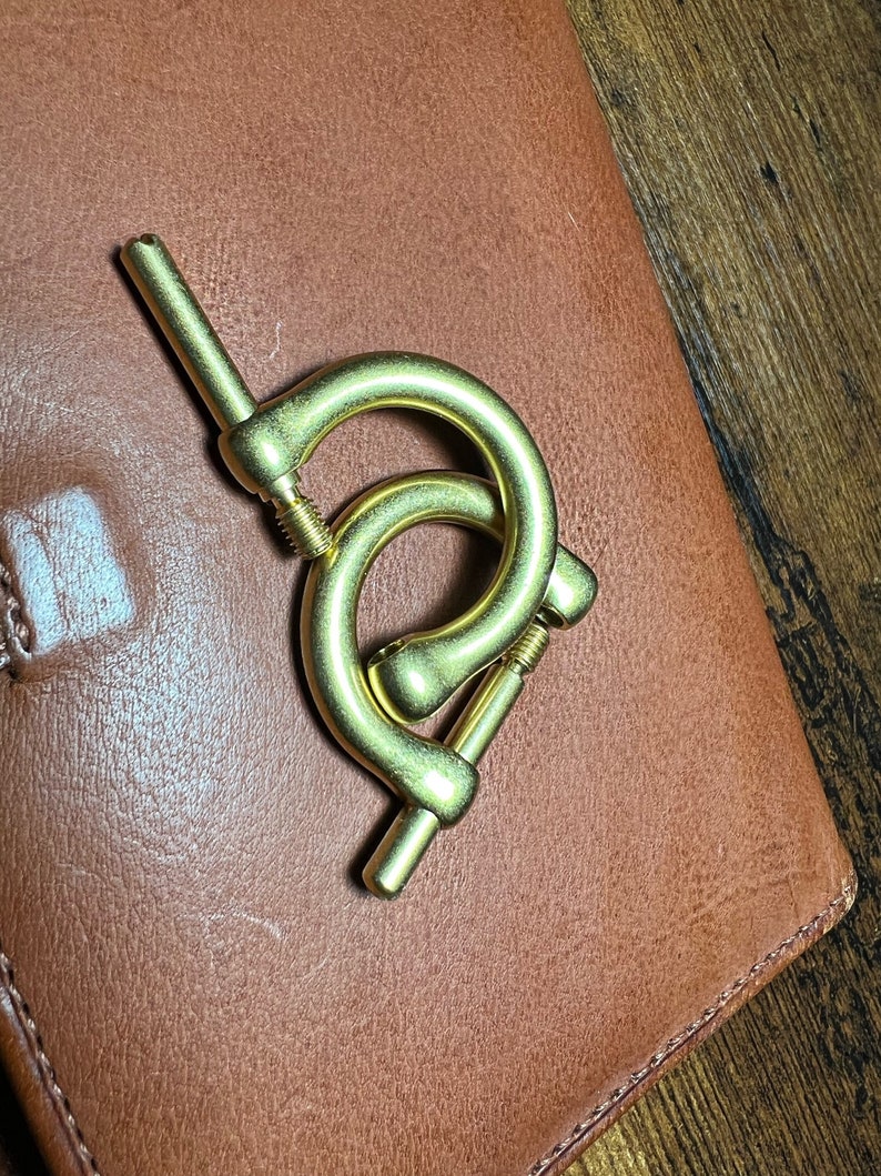 May include: A brass-colored metal clasp with two curved sections and a screw mechanism. The clasp is positioned on a brown leather surface, with a wooden surface visible in the background.