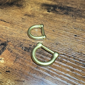 May include: Two brass-colored D-rings are displayed on a wooden surface. The D-rings have a rounded shape with a straight bar connecting the two curved sides. The metal has a shiny, reflective surface.