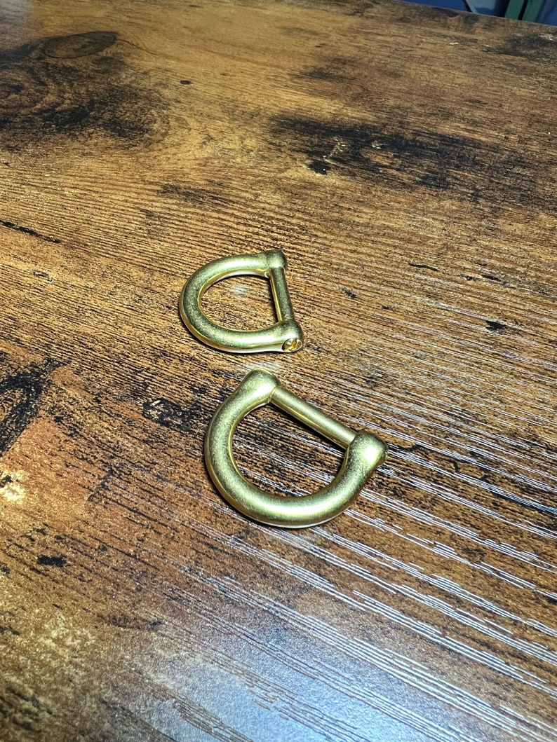 May include: Two brass-colored D-rings are displayed on a wooden surface. The D-rings have a rounded shape with a straight bar connecting the two curved sides. The metal has a shiny, reflective surface.