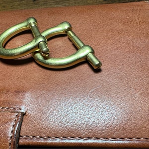 May include: A pair of gold-colored metal shackles, interlocked, resting on a brown leather surface. The shackles have a curved design with a screw closure. The leather appears to be a wallet or notebook.