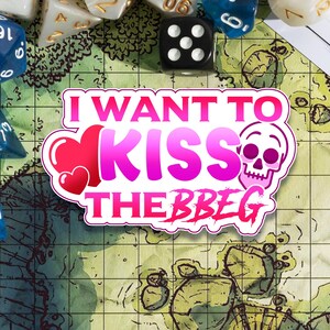 Könnte beinhalten: Ein rosa-weißer Aufkleber mit dem Text "I want to kiss the BBEG" mit einem rosa Herzen und einer Totenkopfgrafik.