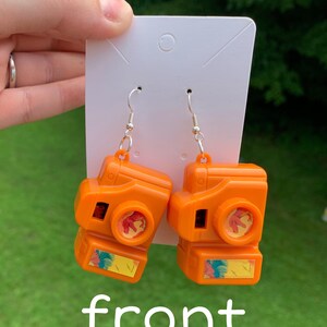 May include: A pair of orange camera-shaped earrings with silver hooks. The earrings have a retro design with a small rectangular image on the bottom and a circular lens. The word "front" is written in white at the bottom of the image.