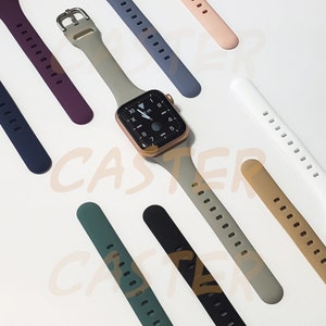 May include: A variety of silicone watch bands in different colors, including gray, white, black, green, blue, and brown. The bands are all designed for Apple Watches and have a textured surface. The bands are arranged in a semi-circle around a gold Apple Watch with a white face.