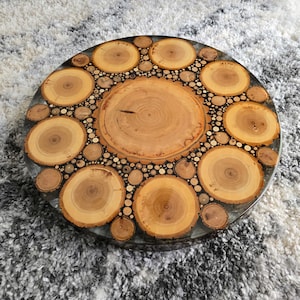 May include: Round wooden table with a unique design. The table features a central large wood slice surrounded by smaller wood slices and pieces, all set in a clear resin. The wood has a natural, light brown color, and the resin has a dark border.