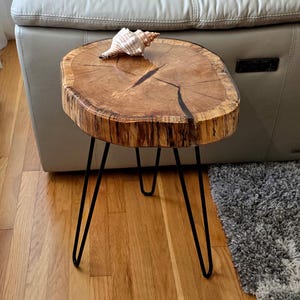 May include: A small, round wooden side table with a natural wood grain finish. The table has a live edge and is supported by three black hairpin legs. A seashell rests on the tabletop. The table measures approximately 18 inches in diameter.