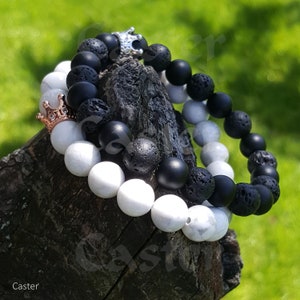 May include: Two bracelets, one with black lava beads and one with white beads, both with a silver crown charm. The bracelets are on a wooden surface.