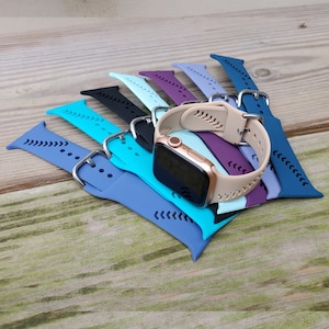 May include: A collection of colorful silicone watch bands for a smartwatch. The bands are in various shades of blue, purple, and beige. The bands have a textured design with small holes for ventilation. The bands are arranged in a stack with a smartwatch face visible at the top.