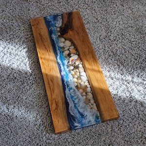 May include: A wooden wall art piece with a blue and white resin wave design. The wave is filled with seashells and is set against a natural wood background.