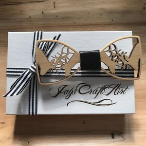 May include: A wooden bow tie featuring a deer silhouette design. The bow tie is presented in a white box with a black and white striped ribbon and the text "Jays Craft Art" printed on the box.