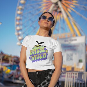 Puede incluir: Top blanco corto con el texto "Just your basic witch" en una fuente retro colorida. El texto está rodeado de un círculo morado con un contorno amarillo y dos murciélagos negros.