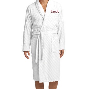 May include: A white, textured bathrobe with a shawl collar and a waist tie. The word "Jacob" is embroidered on the chest. The robe has pockets and a knee-length hemline. The person is standing barefoot.