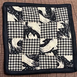 May include: A square quilted potholder featuring a checkerboard pattern of black and white houndstooth fabric alternating with a black fabric printed with white ghosts, stars, and crescent moons, all finished with a solid black fabric border.