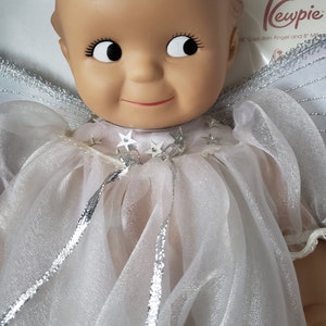 May include: A Kewpie doll dressed as an angel, featuring a smiling face with large eyes and eyelashes. The doll wears a white, sheer gown with silver trim and star accents. The doll has white wings and is approximately 16 inches tall.