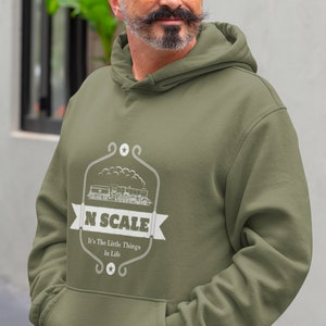 Puede incluir: Una sudadera con capucha verde con un gr&aacute;fico blanco de un tren y el texto "N SCALE It's The Little Things In Life."