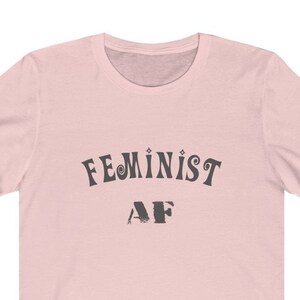 Op de afbeelding: Een roze T-shirt met de tekst "FEMINIST AF" in zwart gedrukt.