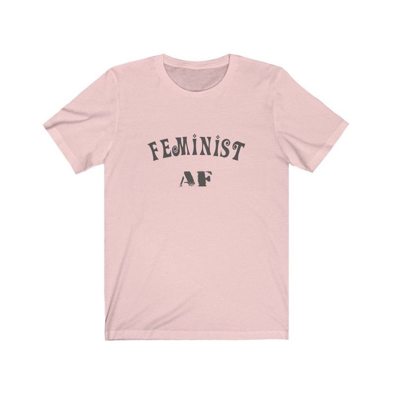 Op de afbeelding: Een roze T-shirt met de tekst "FEMINIST AF" in zwart gedrukt.