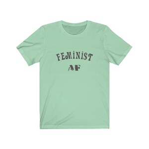 Op de afbeelding: Een lichtgroen T-shirt met de tekst "FEMINIST AF" in zwart.