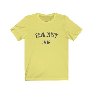Op de afbeelding: Een geel T-shirt met korte mouwen met de tekst "FEMINIST AF" in het zwart.