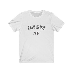 Op de afbeelding: Wit T-shirt met zwarte tekst die "FEMINIST AF" luidt in een retro-lettertype.
