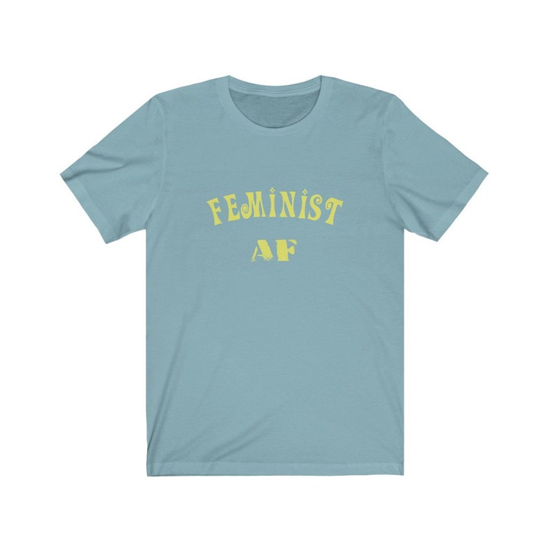 Op de afbeelding: Een lichtblauw T-shirt met de tekst "FEMINIST AF" in geel.