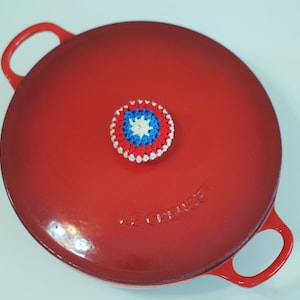 May include: Red enameled cast iron pot with a crocheted pot holder in the shape of Captain America's shield. The pot holder is red, white, and blue.