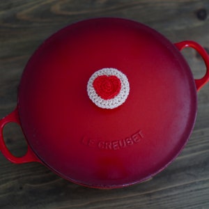 May include: Red enameled cast iron pot with a white crocheted heart-shaped knob on the lid. The pot has the text "Le Creuset" embossed on the lid.