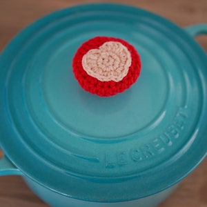 May include: A turquoise blue enameled cast iron pot with a red and white crocheted heart-shaped knob on the lid. The lid has the text "Le Creuset" embossed on it.