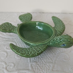 May include: A green ceramic turtle-shaped dish with a textured surface. The dish has a central bowl and four flipper-like extensions. The turtle's head has blue eyes. The dish is designed for decorative or functional use.