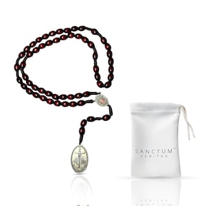 May include: A dark red rosary with oval beads and a silver-toned crucifix pendant. The rosary is displayed next to a white drawstring pouch with the text "SANCTUM VERITAS" printed on it.